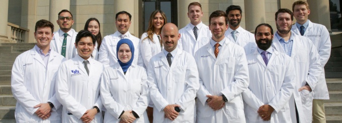 Fourteen smiling residents in white coats in front of a stone building. 