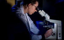 A student researcher in a lab coat peers through a microscope. 