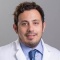 One smiling resident in white coat and blue shirt and tie in front of gray back drop. 