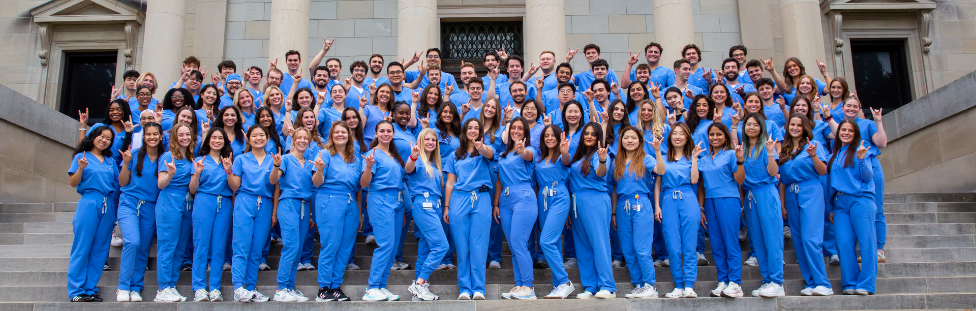 The Class of 2028, clad in blue scrubs, poses on the steps of Abbott Library with their horns up. 