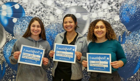 Three dental students in front of a background holding up Match Day signs. 