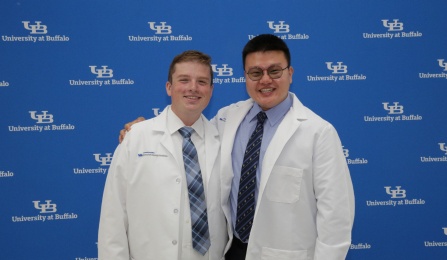 Two dental students in lab coats in front of the UB School of Dental Medicine logo backdrop. One student has their arm around the other. 