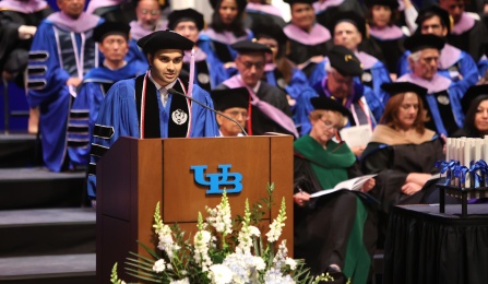 A student dressed in graduation attire standing behind a podium giving a speech. 