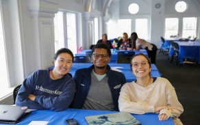 Three dental students sitting behind a table smiling. They have laptops in front of them on the table. 