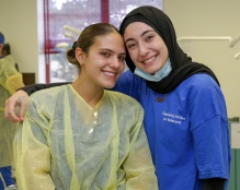 Two dental students in the clinic. One student is wearing a dentist gown with the other student having an arm around her. 