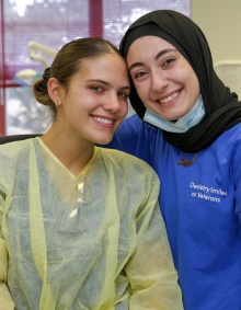 Two dental students in the clinic. One student is wearing a dentist gown with the other student having an arm around her. 