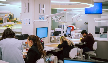 Three dental students sitting behind a table smiling. They have laptops in front of them on the table. 