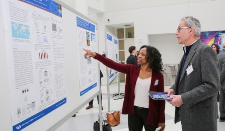 Photo of student pointing at poster presentation with faculty looking on. 