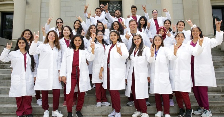 Dental students outside library steps in their white coats holding up their hands. 