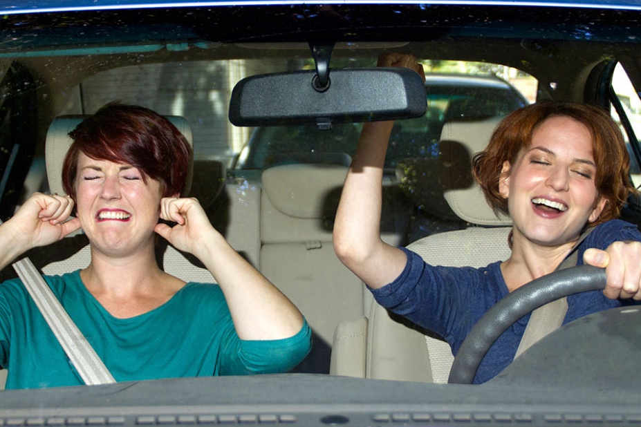 A young woman sings as she drives while another young woman hold her ears with a pained look on her face. 