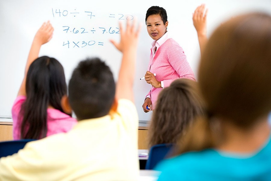 A teacher stands in front of a dry erase board containing math equations, while students raise their hands to answer a question.