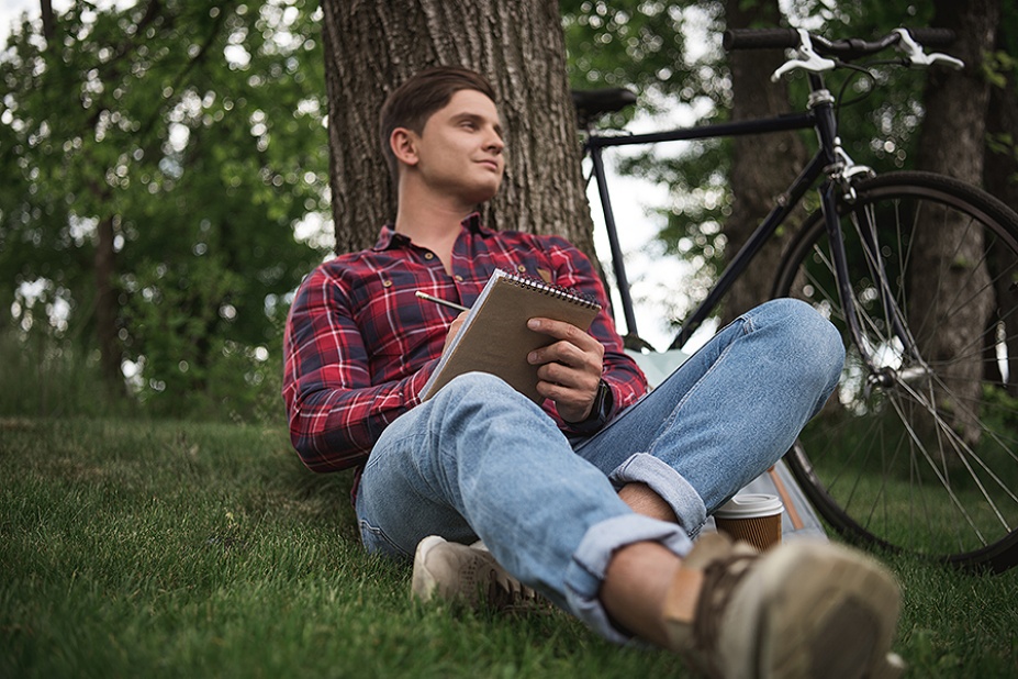 A young man sits in front of a tree with a writing pad in his hand and a bicycle in the background. 