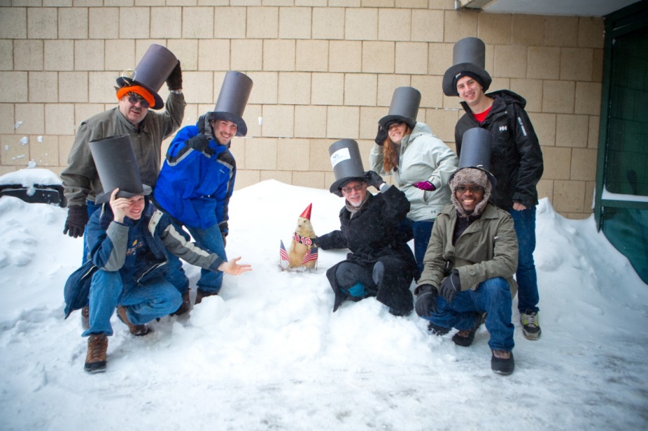 People with paper tophats posing with a taxidermied groundhog in the snow. 