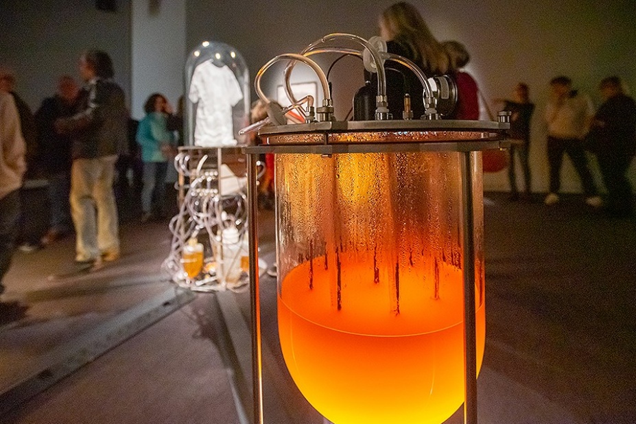 An art gallery exhibition featuring a fermenter filled with orange liquid containing bacteria and an enclosed t-shirt behind it. People fill the room. 