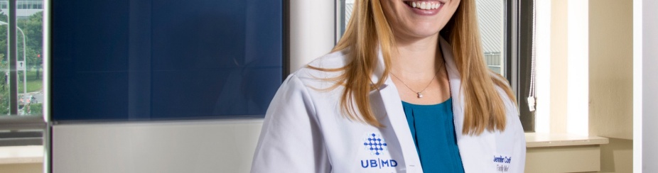 Jennifer Corliss, MD, standing, wearing white coat in front of window. 