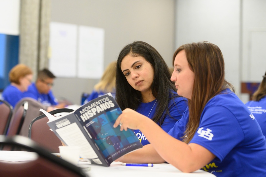 Two people sitting next to each other in blue t-shirts read a book. Other people performing similar tasks are in the background. 