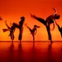 Six dancers kick their legs into the air in front of a orange sunset-like background on the CFA mainstage. 