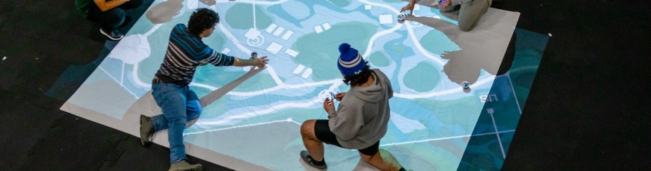 Overhead picture of four students plotting drone flying points on a large map projected on the floor on a lab.